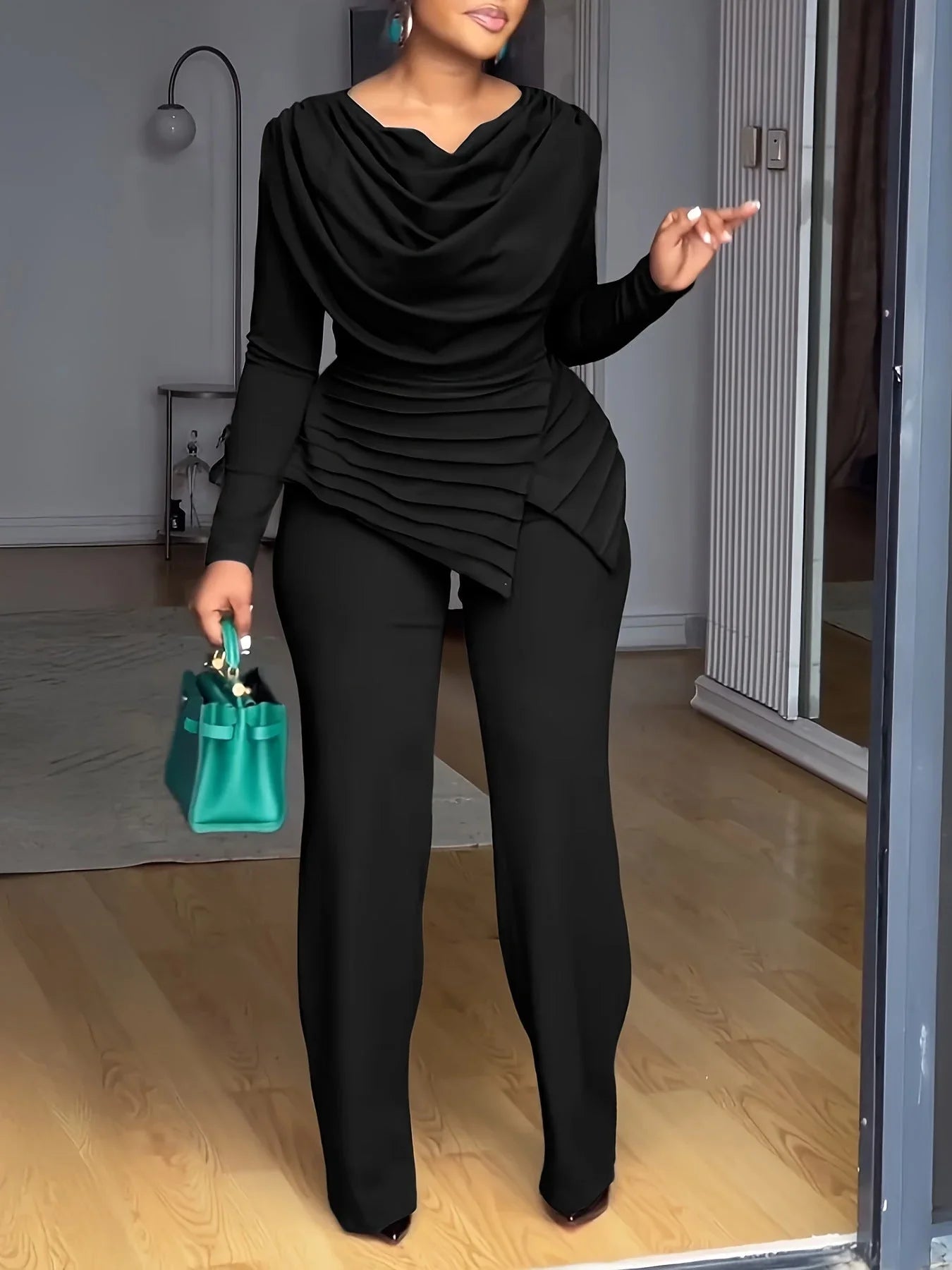 Femme portant un ensemble pantalon noir élégant à col drapé et sac à main vert, intérieur moderne.