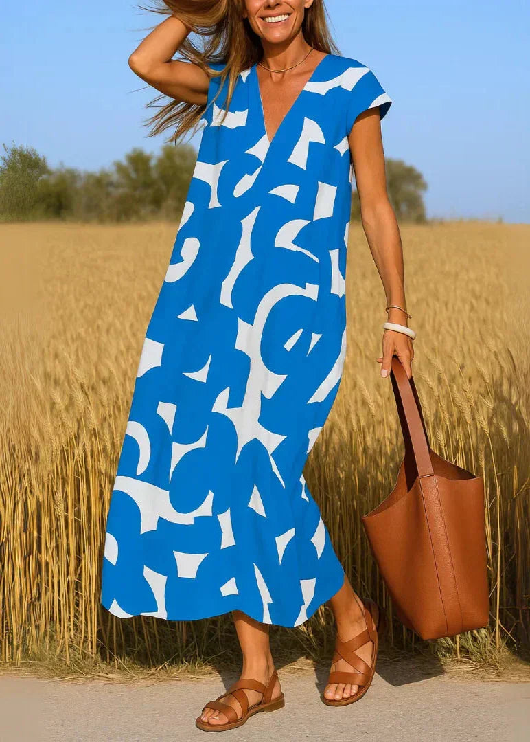Femme en robe longue bleue à motifs blancs, sandales marron, tenant un sac dans un champ