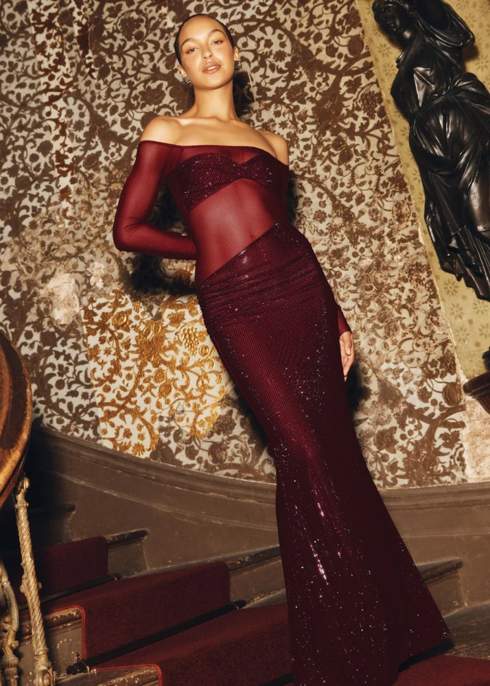 Femme portant une robe longue bordeaux à manches longues transparentes, dans un escalier élégant.