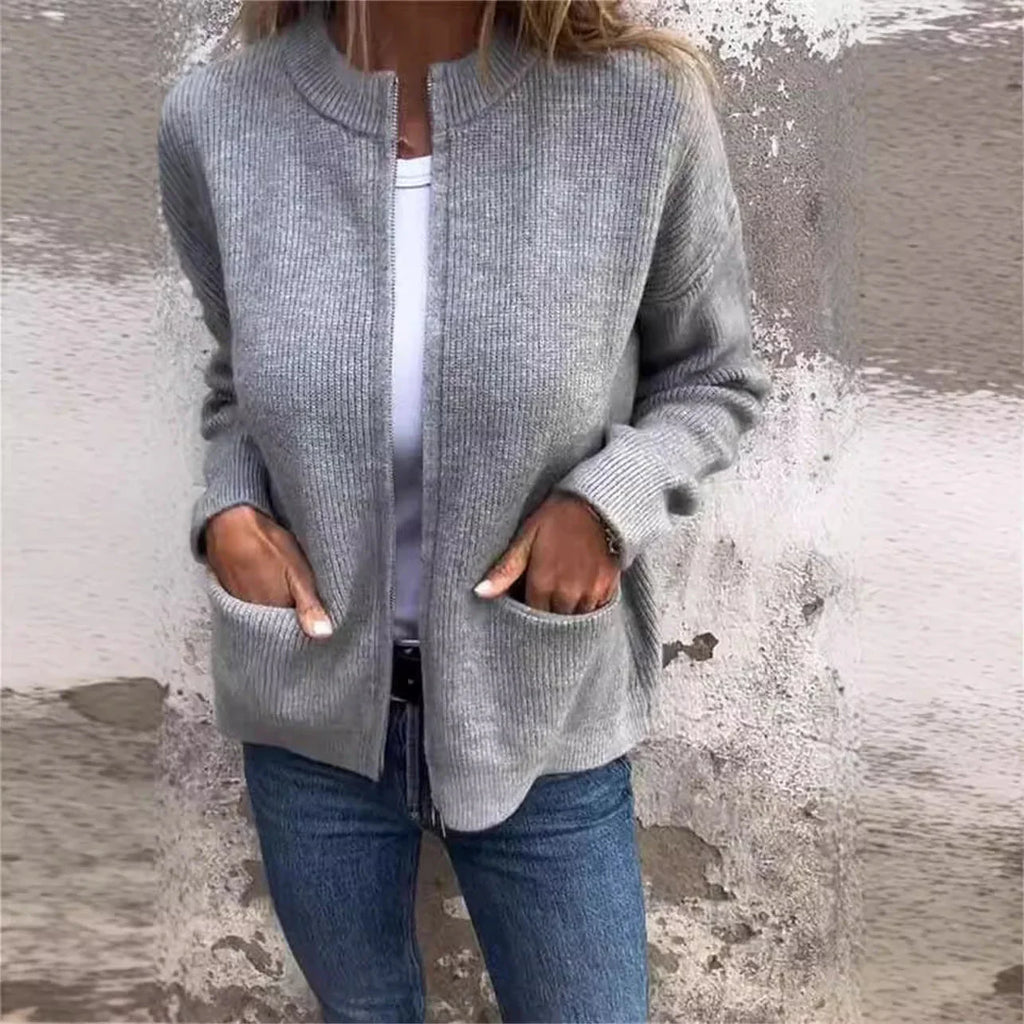 Femme portant un cardigan gris en maille avec poches, t-shirt blanc et jean bleu devant un mur texturé