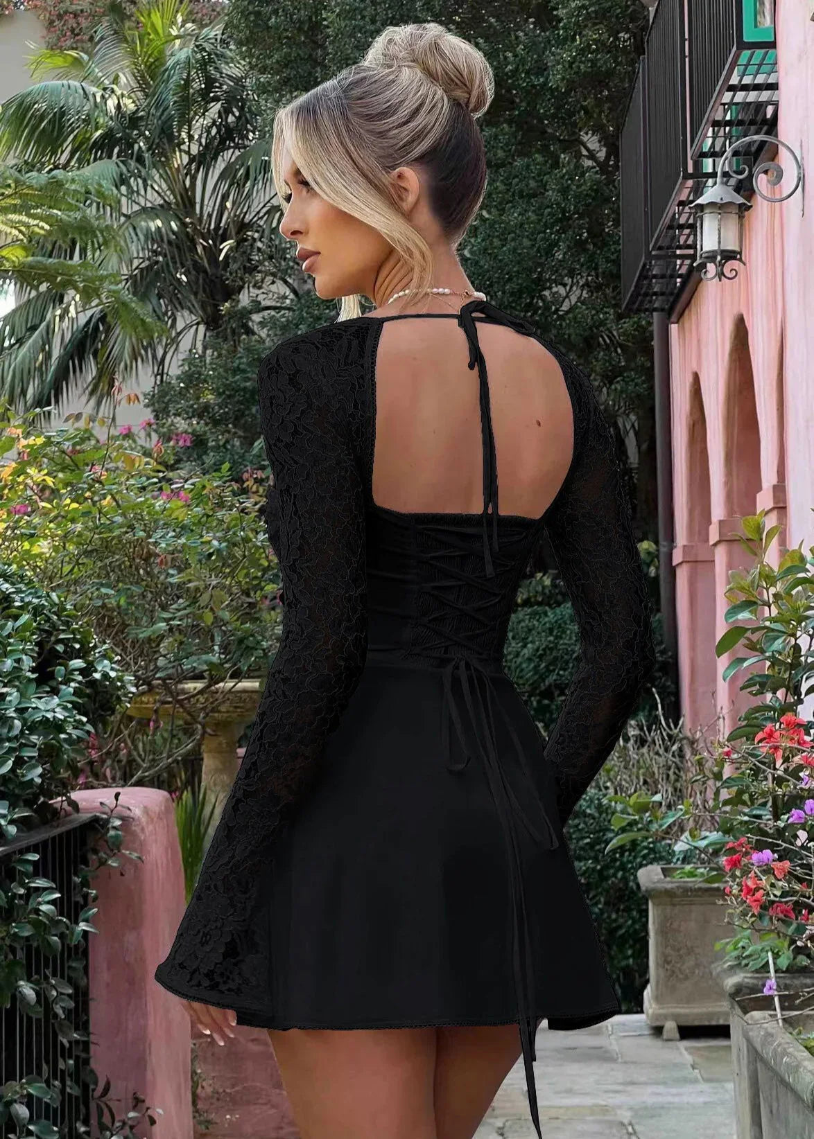 Femme de dos portant une robe noire à manches longues en dentelle, dos nu, en jardin fleuri.